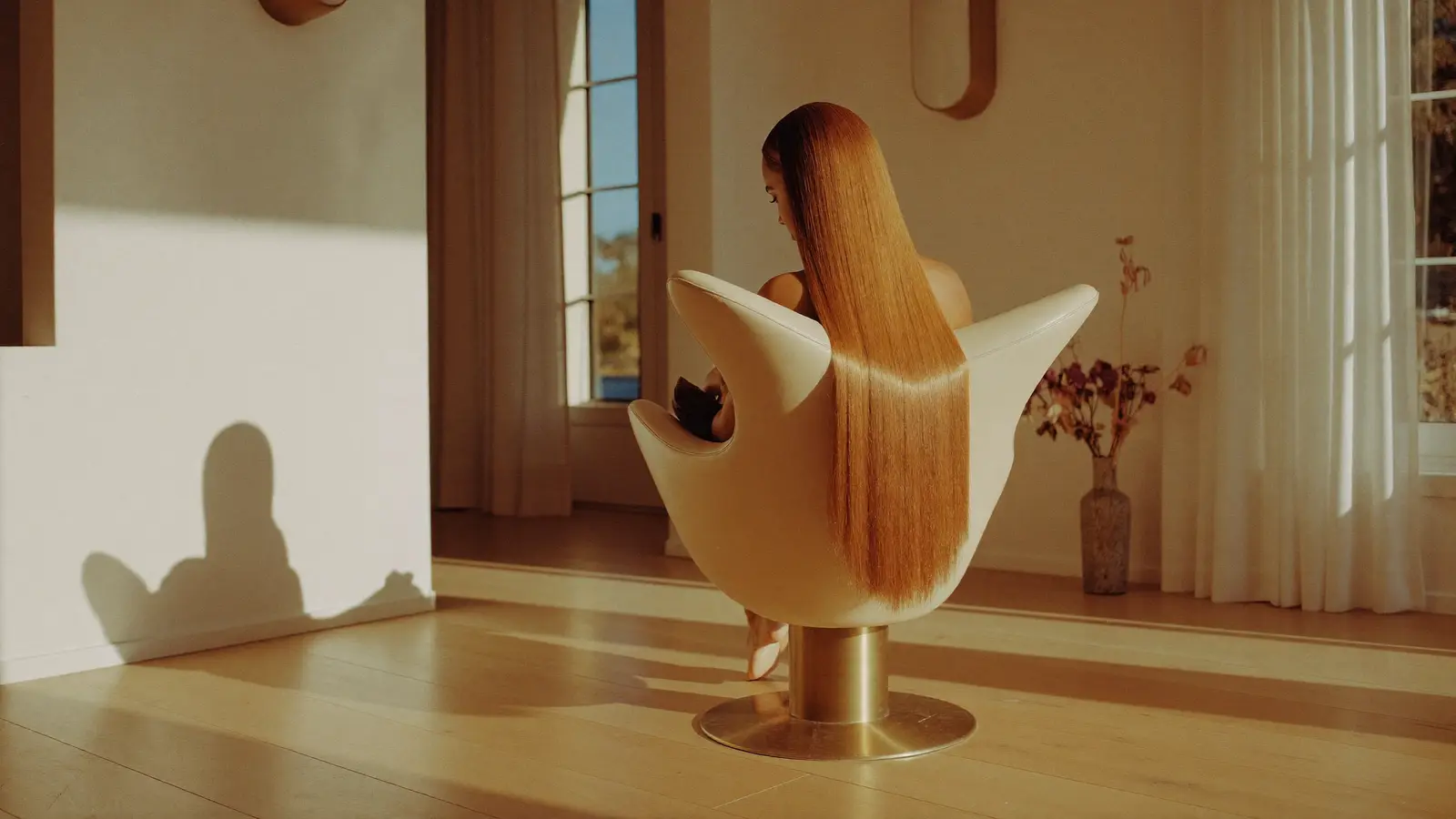 A client with long copper hair seated in a sculptural cream salon chair in afternoon light